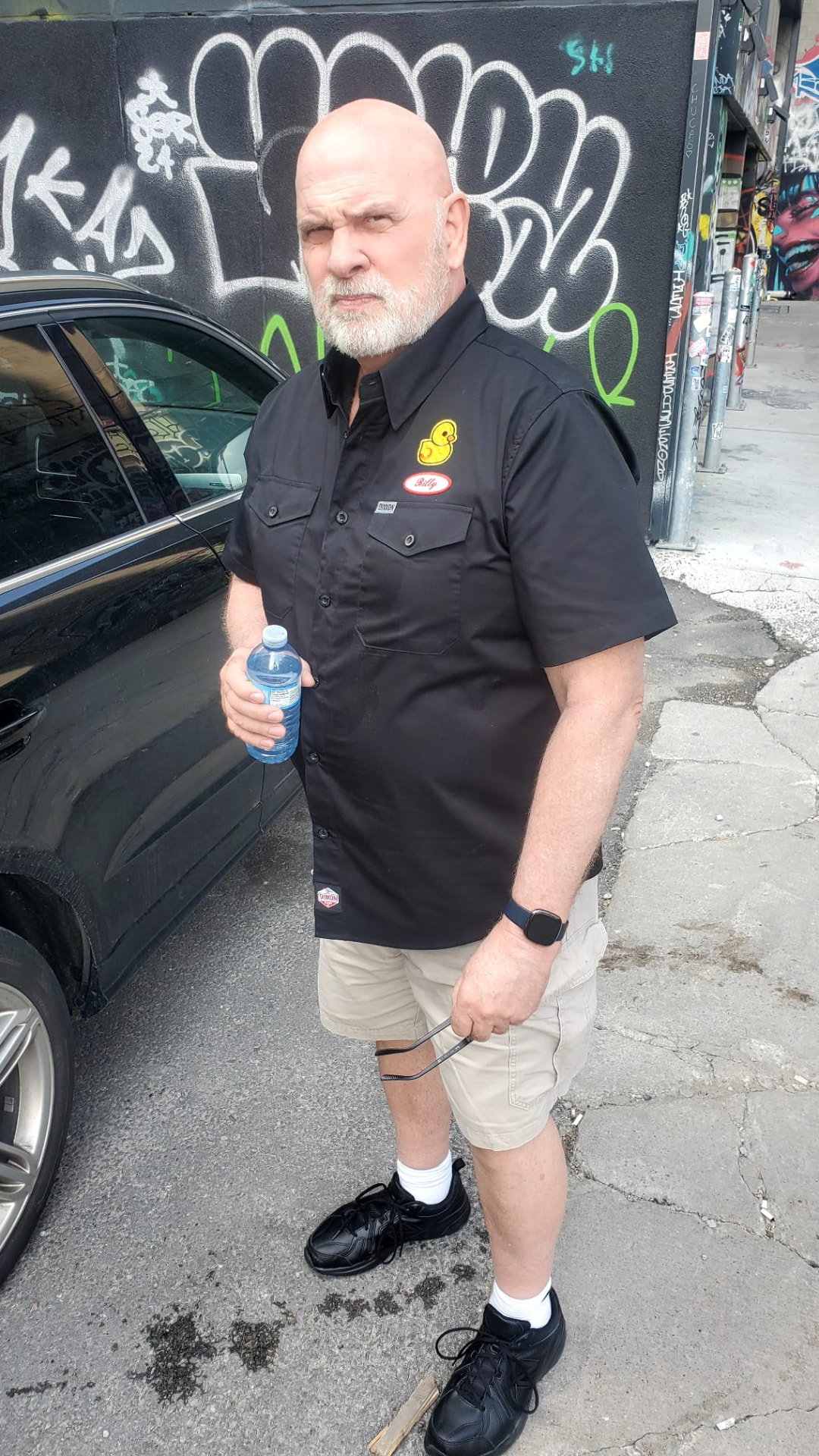 Billy Baker as Bossy Billy beside a black car and graffiti wall in Chain Link, 2025
